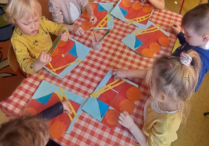 dzieci przy stolikach do wozów strażackich na dachu przyklejają żółtą drabinę składająca się z długich płaskich prostokątów oraz czterech małych prostokątów troczących szczeble