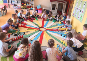 dzieci na wiatraku matematycznym na wstążce, której siedzą układają odpowiednia ilość plastikowych kubeczków wg liczby na wstążce