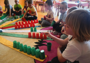 dzieci na wiatraku matematycznym na wstążce, której siedzą układają odpowiednia ilość plastikowych kubeczków wg liczby na wstążce