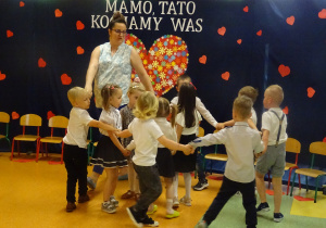 dzieci tańczą na tle dekoracji "Mamo, tato kocham Was" wraz z panią w dwóch kółeczkach