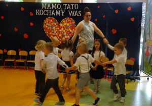 dzieci tańczą na tle dekoracji "Mamo, tato kocham Was" wraz z panią w dwóch kółeczkach
