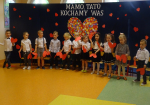 dzieci tańczą z papierowymi sercami na tle dekoracji "Mamo, tato kocham Was"