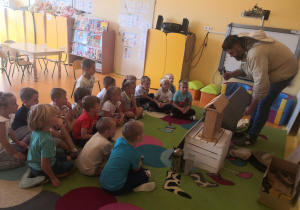 Dzieci siedzą na dywanie w kole i słuchają prezentacji Pana pszczelarza.