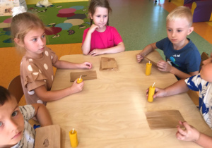 Dzieci siedzą przy stoliku z wykonanymi świecami z wosku pszczelego.