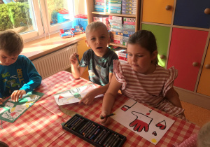 Dzieci siedzą przy stoliku i malują pastelami rysunki na temat Przedszkolaki dla Powstańców.