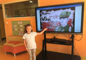 Dziewczynka wskazuje na monitor interaktywny, na którym widać żołnierza, flagę Polski, orła, rok 1918 oraz napis Powstanie Wielkopolskie.