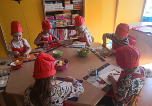 Przedszkolaki siedzą przy stoliku i kroją owoce.