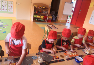 Przedszkolaki siedzą w fartuszkach i czapkach kucharskich przy stoliku i kroją owoce.