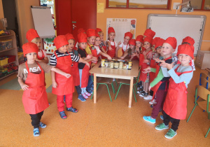 Dzieci stoją przy stoliku ubrane w fartuszki i czapki kucharza, wskazują na przetwory stojące na stoliku.