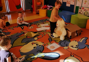 Dzieci siedzą na dywanie, chłopiec ustawia misia przed pudełkiem.