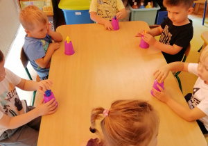 Dzieci siedzą przy stoliku z kubkami plastikowymi odwróconymi do góry nogami, na kubkach stawiają małą figurkę..
