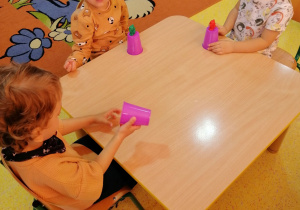 Przedszkolaki siedzą przy stoliku, kubki plastikowe stoją na stoliku, na kubkach dzieci położyły małe figurki.