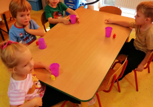 Dzieci siedzą przy stoliku, kubki plastikowe stoją na stoliku, małe figurki są ustawione przed kubkami.