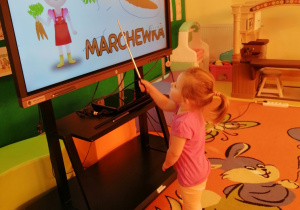 Dziewczynka wskazuje wskaźnikiem marchewkę na monitorze interaktywnym.
