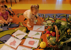 Dzieci siedzą na dywanie, rozłożone są na nim ilustracje z warzywami oraz stoi na nim kosz wypełniony warzywami. Dziewczynka sięga po ilustracje z marchewką.