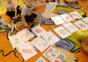 Dzieci siedzą na dywanie, rozłożone są na nim ilustracje z warzywami, chłopiec trzyma w ręku jedną z ilustracji.