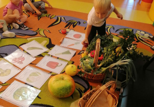 Dzieci siedzą na dywanie, rozłożone są na nim ilustracje z warzywami oraz stoi na nim kosz wypełniony warzywami. Chłopiec stoi przy koszu z warzywami i trzyma w ręku buraka.