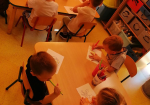 Przedszkolaki siedzą przy stolikach i kolorują obrazki warzyw.