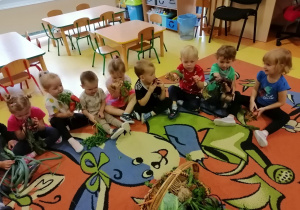 Dzieci siedzą na dywanie, przed nimi stoi kosz z warzywami. Każde dziecko trzyma jedno warzywo.