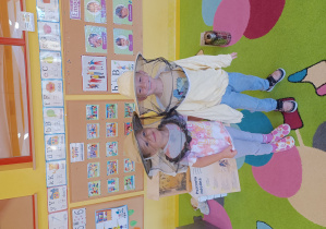 Dziewczynka i chłopiec pozują w strojach pszczelarza, obok dziewczynki stoi szklany ul.