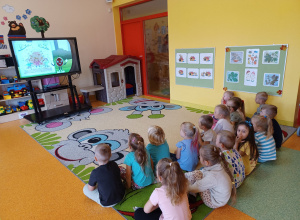 Dzieci siedzą na dywanie i oglądają prezentacje na monitorze interaktywnym.