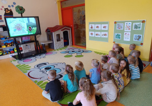 Dzieci siedzą na dywanie i oglądają prezentacje na monitorze interaktywnym.