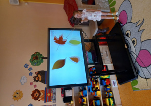 Dziewczynka wskazuje za pomocą wskaźnika na liścia wyświetlonego na monitorze interaktywnym.