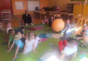 Dzieci wspólnie z panem fizjoterapeutą wykonują ćwiczenia na dywanie przy użyciu dużej piłki.