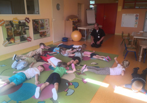 Dzieci wspólnie z panem fizjoterapeutą wykonują ćwiczenia na dywanie; dzieci leżą na brzuchu i wyciągają ręce na boki.