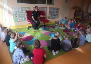 Dzieci siedzą na dywanie i rozmawiają z panią policjantką. Pani policjantka siedzi na krześle.