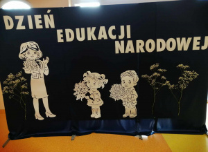Zdjęcie przedstawia dekorację, na granatowym materiale przywieszony jest napis Dzień Edukacji Narodowej i postać nauczycielki z kwiatami, dziewczynki oraz chłopca trzymających kwiaty.