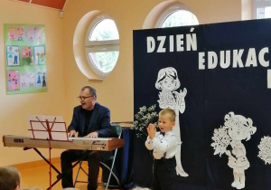 Na pianinie gra Pan Sławomir, śpiewa chłopiec, dzieci siedzą na krzesłach.