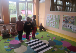 Dzieci siedzą na dywanie, dwie dziewczynki i chłopiec stoją przed pasami trzymając się za ręce.