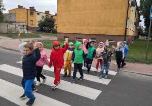 Dzieci przechodzą przez przejście dla pieszych, mają jedną rękę podniesioną do góry.
