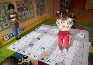 Przedszkolaki siedzą na dywanie, rozłożona jest mata do kodowania, dziewczynka chodzi po macie, chłopiec stoi na dywanie.