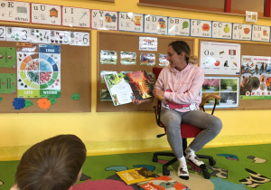 Dzieci siedzą na dywanie, Pani Marta siedzi na fotelu czyta książkę i pokazuje ilustracje.