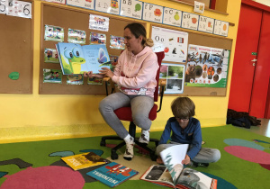 Pani Marta siedzi na fotelu czyta książkę i pokazuje ilustracje, Stasiu siedzi na dywanie i ogląda książkę.