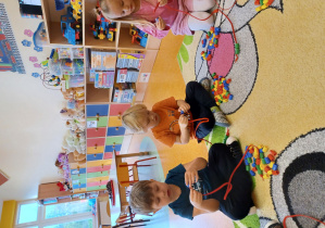 Dzieci siedzą na dywanie i nawlekają kołeczki na sznurki.