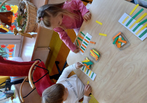 Dzieci siedzą przy stolikach, przyklejają paseczki na kartce według określonego rytmu.