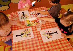 Dzieci siedzą przy stoliku i wykonują prace plastyczną pod tytułem Jesienne drzewo".