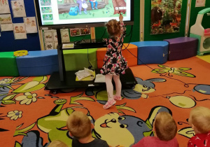 Dzieci siedzą na dywanie, dziewczynka wskazuje na sowę na ekranie interaktynym.