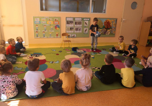 Dzieci siedzą na dywanie, Wojtek stoi na dywanie i trzyma piłkę.