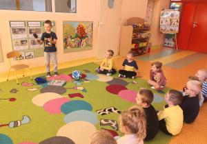 Dzieci siedzą na dywanie, Wojtek stoi na dywanie i pokazuje dzieciom rękawice bramkarza.