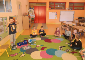 Dzieci siedzą na dywanie, Wojtek stoi na dywanie i pokazuje dzieciom medal.