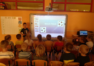 Dzieci siedzą na krzesłach przed tablicą interaktywną, Wojtek stoi przy tablicy, na której wyświetlana jest prezentacja o jego zainteresowaniach.