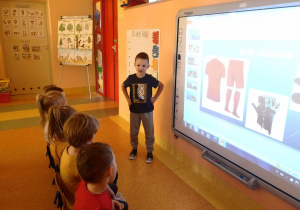 Dzieci siedzą na krzesłach przed tablicą interaktywną, Wojtek stoi przy tablicy i opowiada o tym co wyświetla się na tablicy.