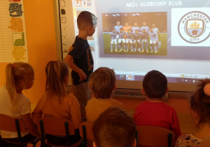 Dzieci siedzą na krzesłach przed tablicą interaktywną, Wojtek stoi przy tablicy i opowiada o tym co wyświetla się na tablicy.