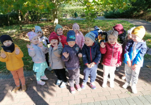 Dzieci pozują do zdjęcia w parku, niektóre przedszkolaki machają ręką.