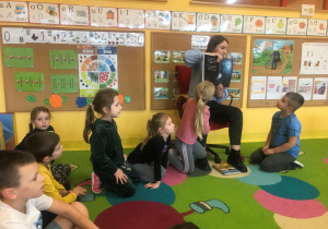 Dzieci siedzą na dywanie, mama Kaliny pokazuje dzieciom ilustracje z bajki.