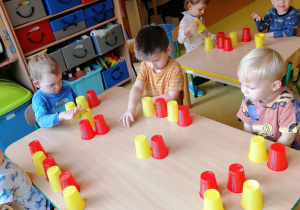 Dzieci siedzą przy stoliku i układają rytm z kolorowych kubeczków.